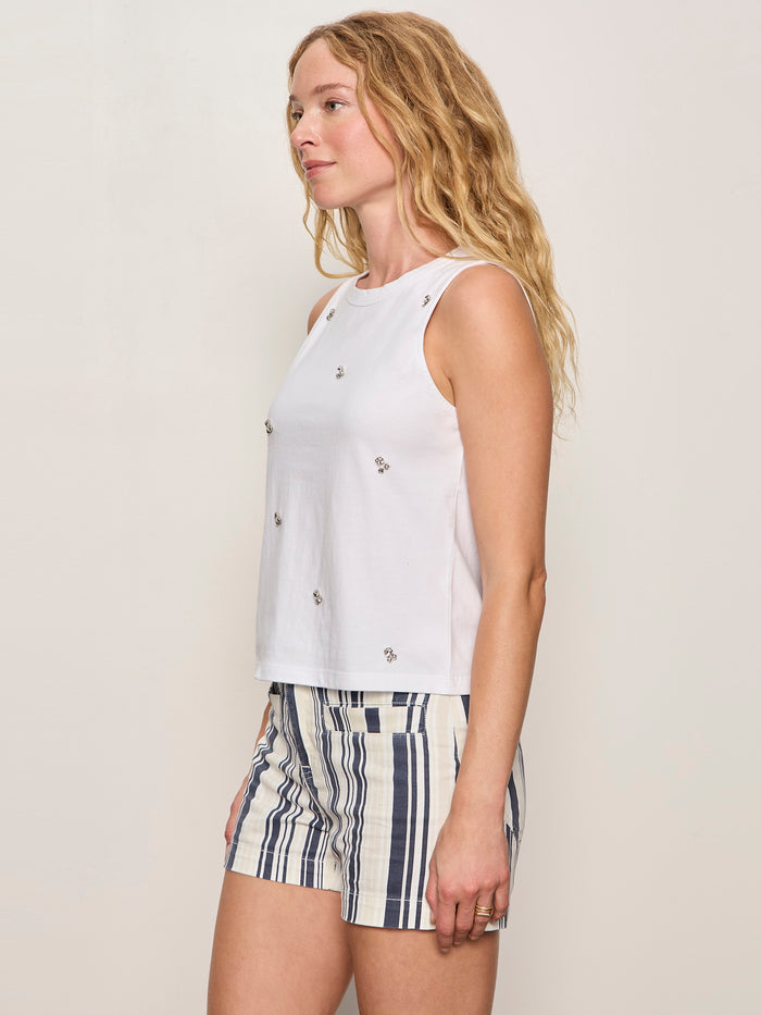 A woman with long wavy blonde hair wears the Sanctuary Clothing campari embellished tank in white. She stands in profile against a plain light background, paired with high-waisted blue, black, and cream striped shorts.