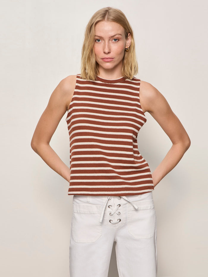 A blonde woman wears the Sanctuary Clothing campari tank clay stripe, a sleeveless brown and white striped top, paired with white lace-up waist pants, standing against a plain light background with her hands behind her back.