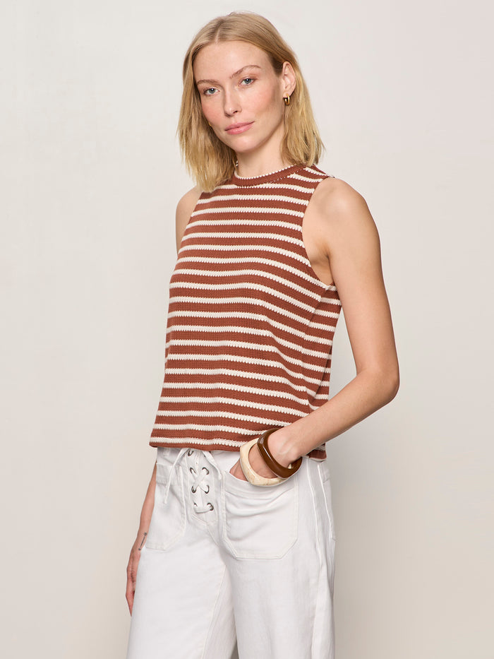 A woman with blonde hair models the Sanctuary Clothing campari tank clay stripe, a sleeveless brown and white striped top, paired with white pants. She stands against a plain light background, gazing at the camera with a relaxed expression.