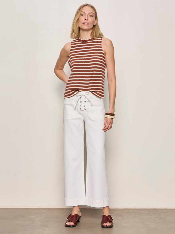 A woman stands against a plain wall, wearing the Sanctuary Clothing campari tank clay stripe top, high-waisted white wide-leg pants with lace-up detail, brown sandals, and gold bracelets.