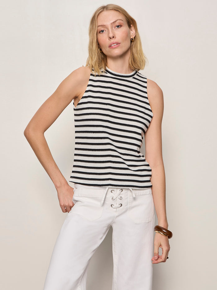 A woman with blonde hair wears the Sanctuary Clothing campari tank macrame stripe top and white lace-up pants, standing against a plain light background with a brown bangle on her wrist.