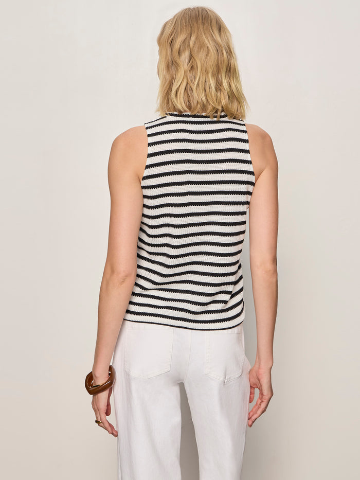 A person with wavy blonde hair models Sanctuary Clothing's campari tank macrame stripe, facing away in a black and white striped sleeveless top and white pants against a plain light background.