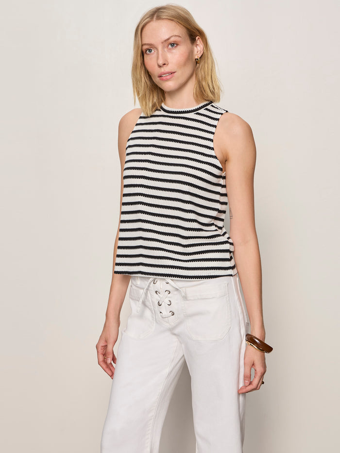 A woman models the Sanctuary Clothing campari tank macrame stripe, a sleeveless black-and-white striped top, paired with white lace-up pants against a plain light background, her head slightly tilted and one hand at her side.