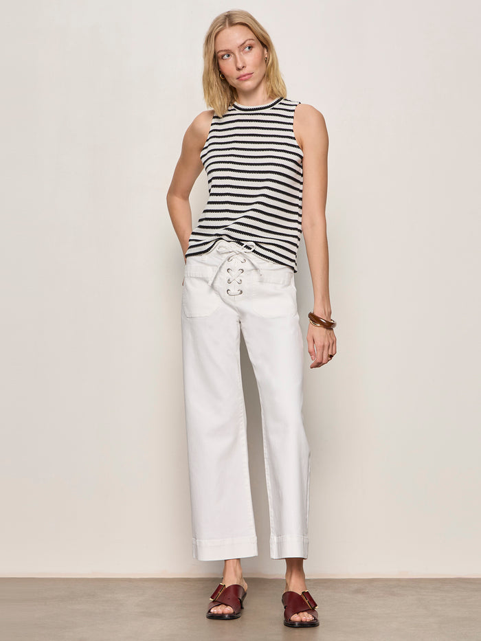 A woman poses against a plain background wearing the Sanctuary Clothing campari tank macrame stripe, white wide-leg lace-up pants, brown sandals, and a few bracelets.