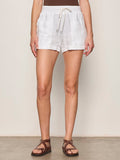 Wearing Sanctuary Clothing's carefree short white, a person poses against a plain light wall and concrete floor, styled with a brown sleeveless top and brown strappy sandals.