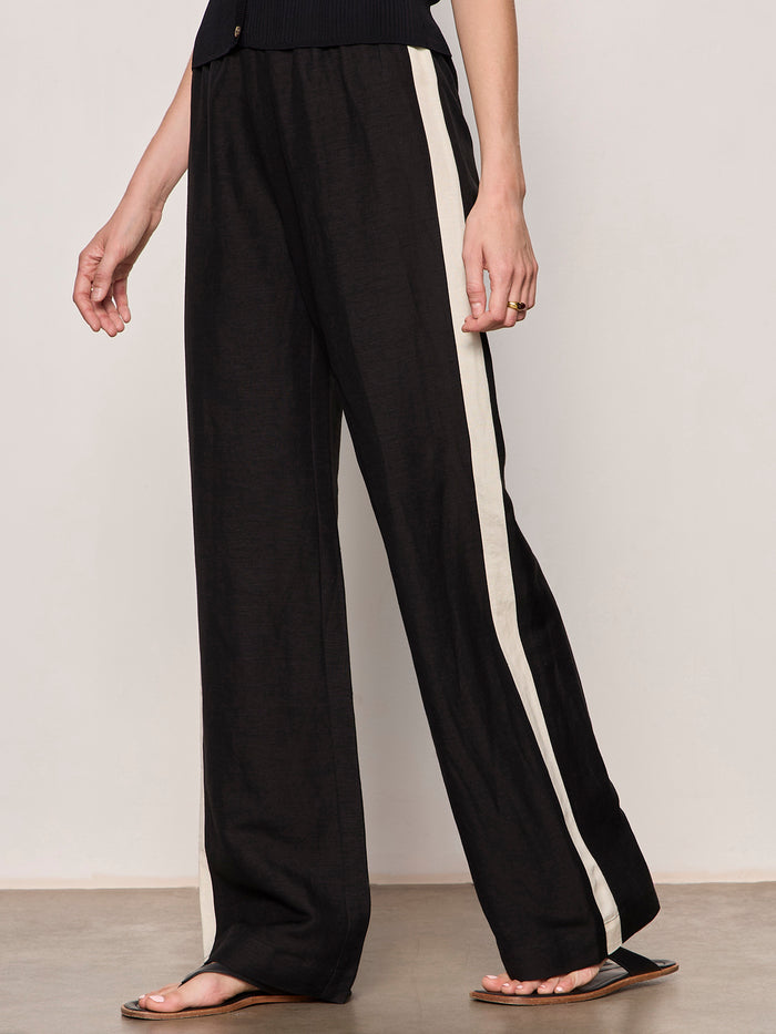 A model wears Sanctuary Clothing’s black casual track pant summer sand with a white side stripe, paired with a black top and brown flat sandals, standing against a plain light background.
