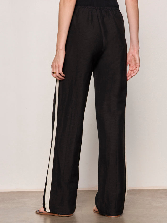 A person stands indoors from the waist down, wearing Sanctuary Clothing's casual track pant black summer sand—loose black pants with a white side stripe, elastic waistband, and flat sandals. The light background is plain.