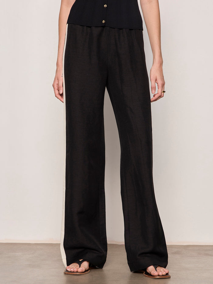 A person wears the Sanctuary Clothing casual track pant in black summer sand with a white side stripe, paired with a black sleeveless top and sandals, standing against a plain background.