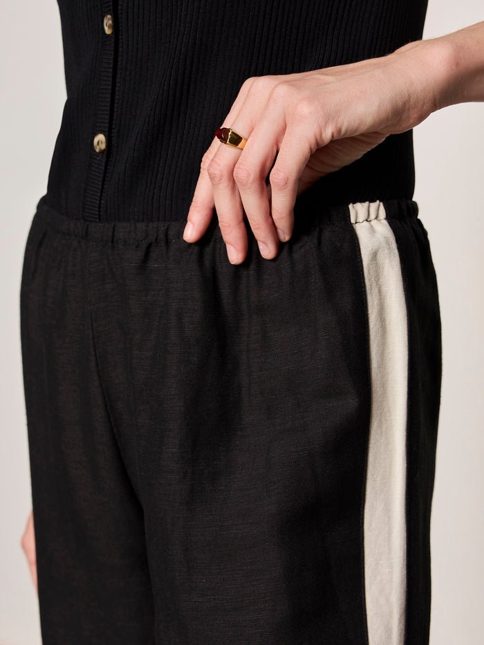 Wearing Sanctuary Clothing's casual track pant black summer sand, a person with their hand on their hip showcases a gold ring, paired with a black ribbed button-up top featuring a white side stripe.