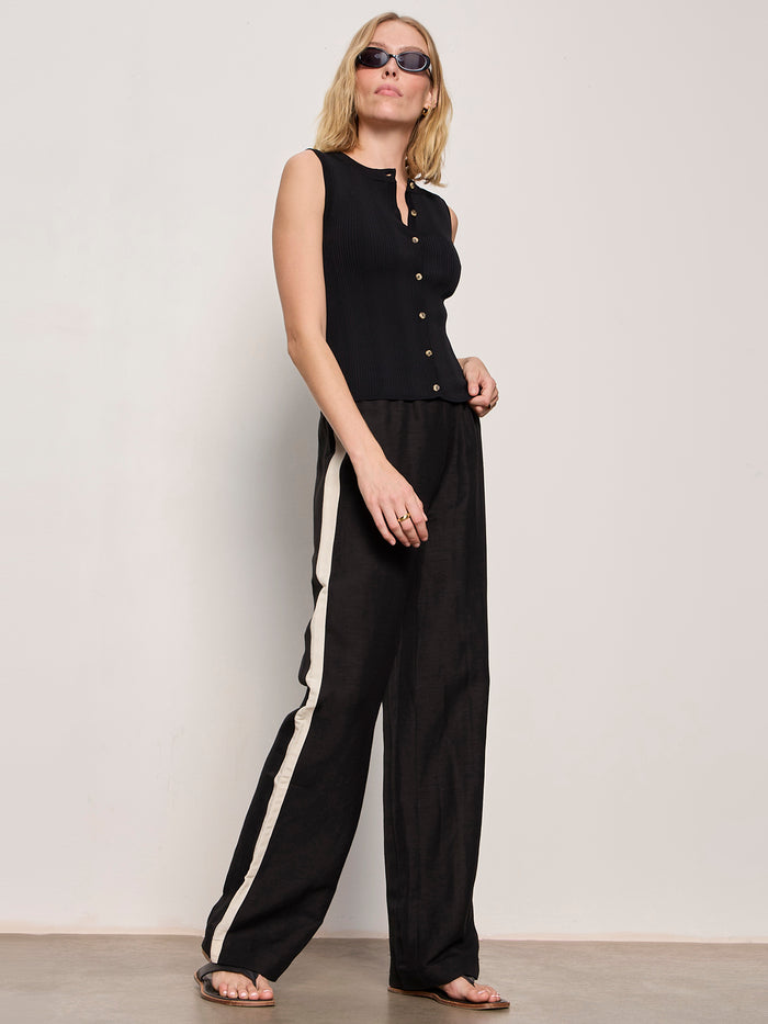 A woman in Sanctuary Clothing's casual track pant black summer sand poses confidently against a plain light wall, pairing the pants with sunglasses and a sleeveless black buttoned top.