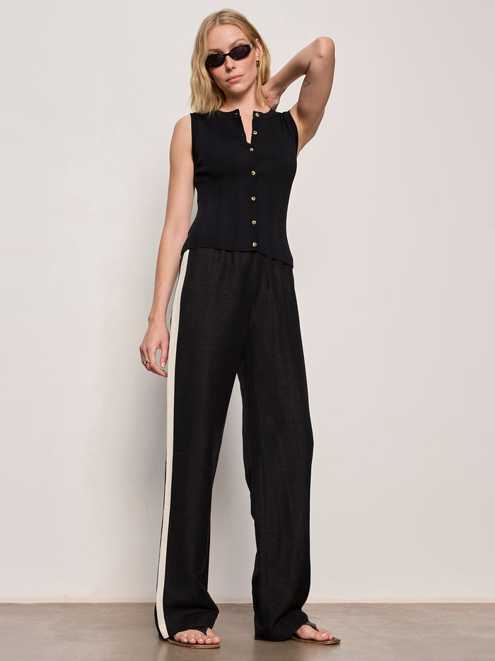 A woman in Sanctuary Clothing's casual track pant black summer sand pairs it with a sleeveless black top and sunglasses, standing against a plain light background with one arm bent and her hand touching her head.