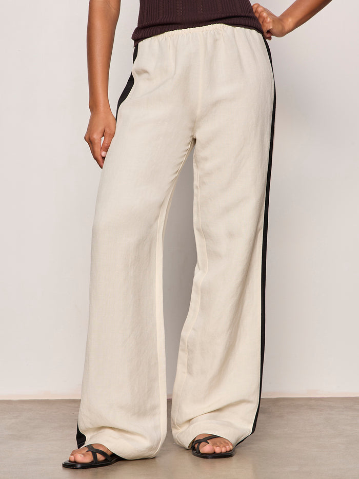 A person models Sanctuary Clothing's casual track pant summer sand black—loose-fitting, cream wide-leg pants with black side stripes—paired with a dark sleeveless top and black sandals, posing against a plain background.
