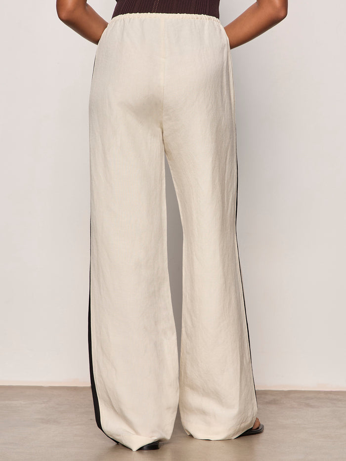 A person wears Sanctuary Clothing's casual track pant summer sand black, featuring loose cream fabric with black side stripes, standing with their back to the camera against a plain background.