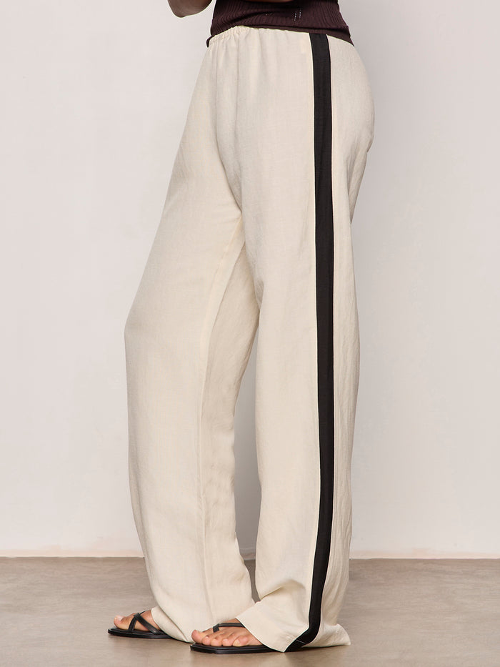 A person models the Sanctuary Clothing casual track pant summer sand black, featuring a relaxed fit and wide black side stripe, standing barefoot on a smooth floor against a plain, light-colored wall.