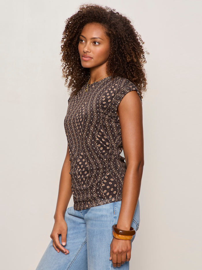 A woman with curly hair stands sideways, wearing the Sanctuary Clothing charisma dolman mesh tee rhythmic, paired with light blue jeans and a brown bangle, against a plain light background.