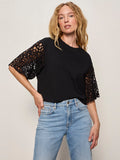 A woman with long, wavy blonde hair stands against a plain background, confidently modeling the Sanctuary Clothing crochet sleeve belle tee in black with lacy short sleeves and light blue jeans, one hand resting on her hip.