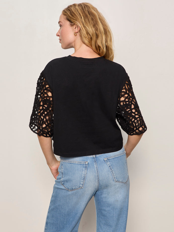 A woman faces away, wearing the Sanctuary Clothing crochet sleeve belle tee in black with intricate lace sleeves and light blue jeans. Her blonde hair is down, hand on hip, set against a plain light background.
