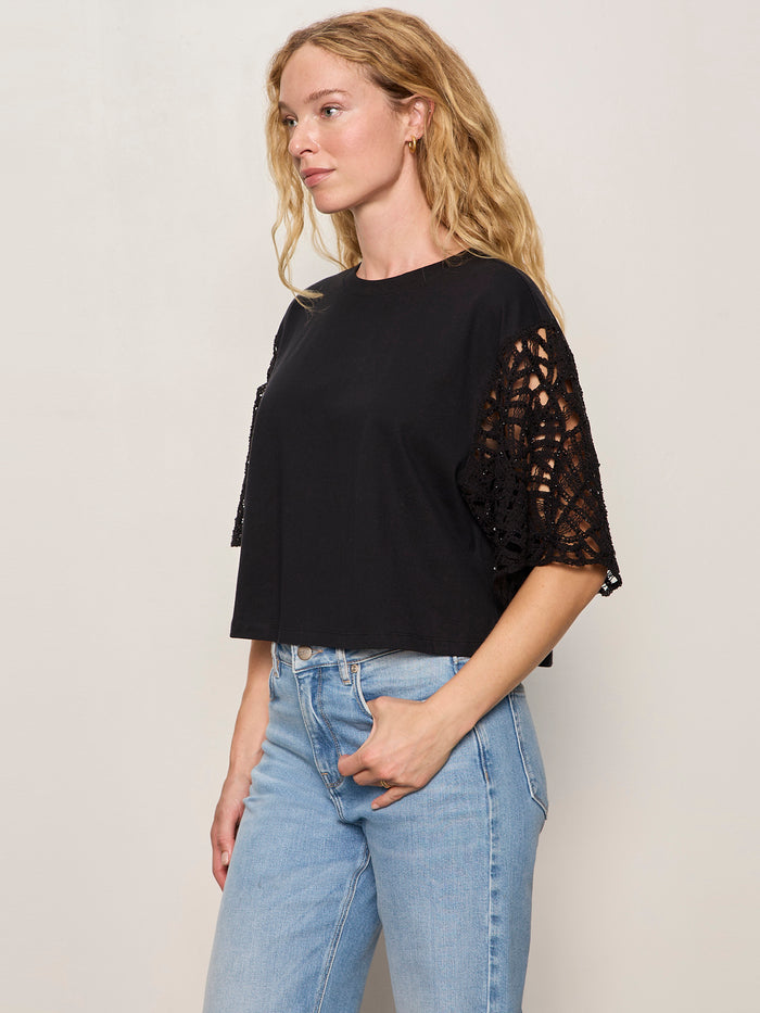 A woman with wavy blonde hair wears the Sanctuary Clothing crochet sleeve belle tee in black and light blue high-waisted jeans, standing against a plain light-colored background.