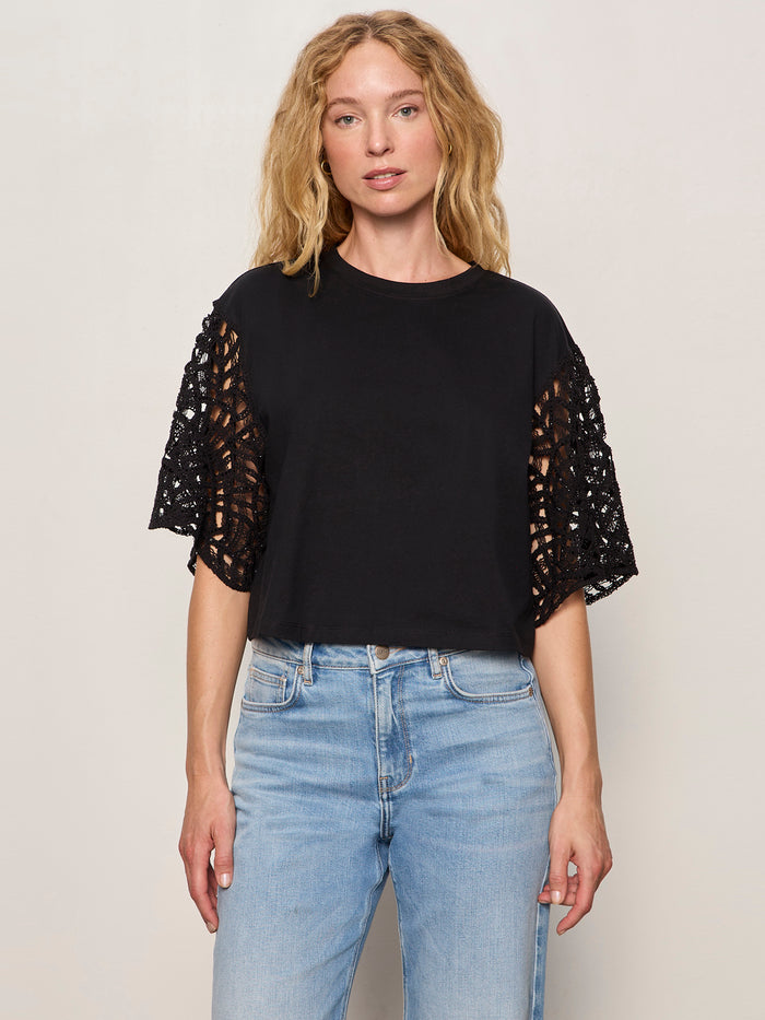 A woman with wavy blonde hair wears the Sanctuary Clothing crochet sleeve belle tee in black with lace cutout sleeves and light blue high-waisted jeans, standing against a plain light-colored background.