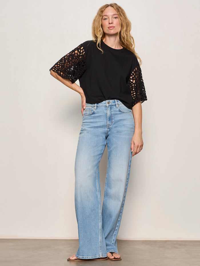 A woman stands against a light-colored wall wearing the Sanctuary Clothing crochet sleeve belle tee in black, paired with light blue high-waisted wide-leg jeans and black sandals.
