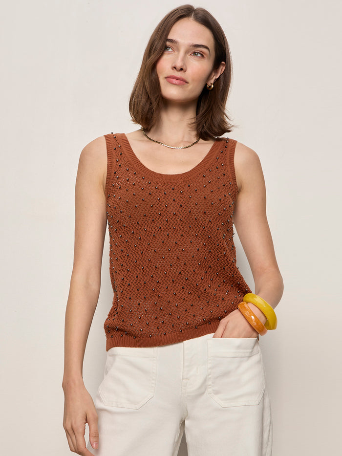 A woman with shoulder-length brown hair wears the Sanctuary Clothing dune studded tank in ancient clay and white pants, accessorized with yellow bracelets and a necklace, standing against a plain beige background.