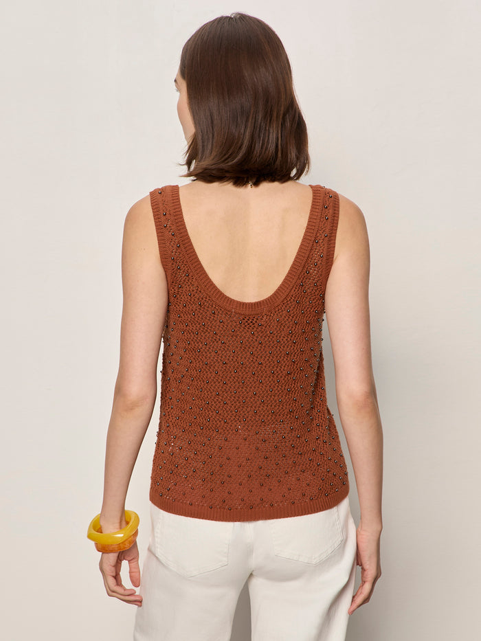 A woman with shoulder-length brown hair is shown from the back wearing the Sanctuary Clothing dune studded tank in ancient clay, paired with white pants and a large yellow bangle, against a plain light background.
