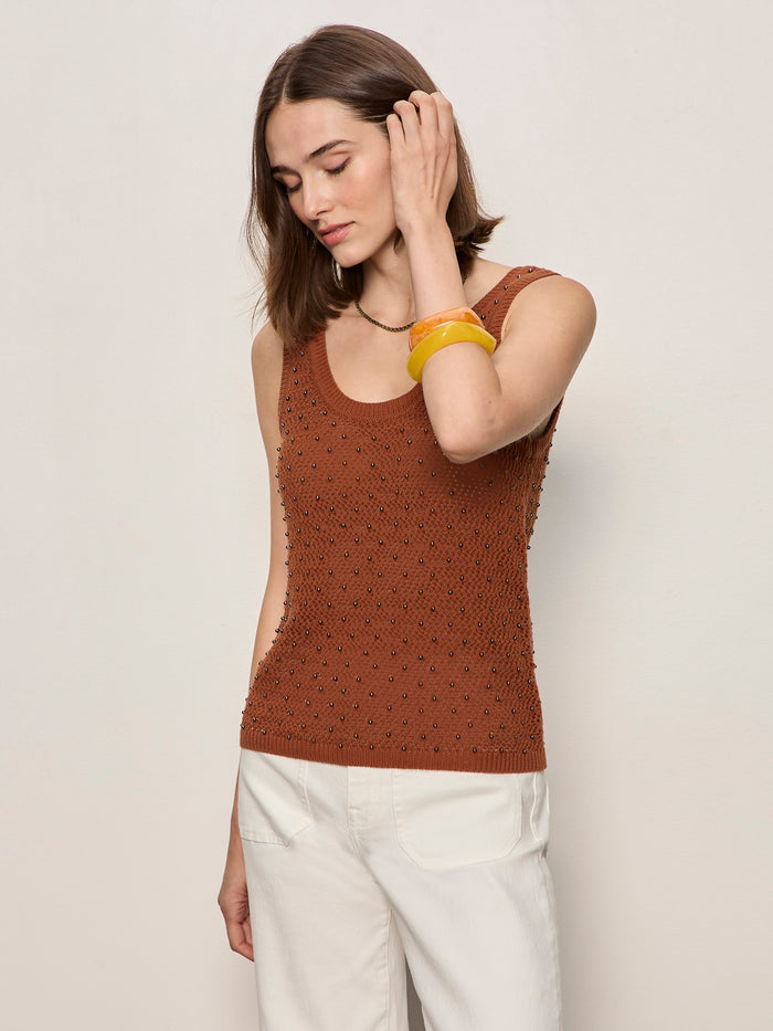 A woman with shoulder-length brown hair wears the Sanctuary Clothing dune studded tank in ancient clay and white pants. She accessorizes with a yellow bangle, gently touching her hair as she looks down, set against a plain light wall.