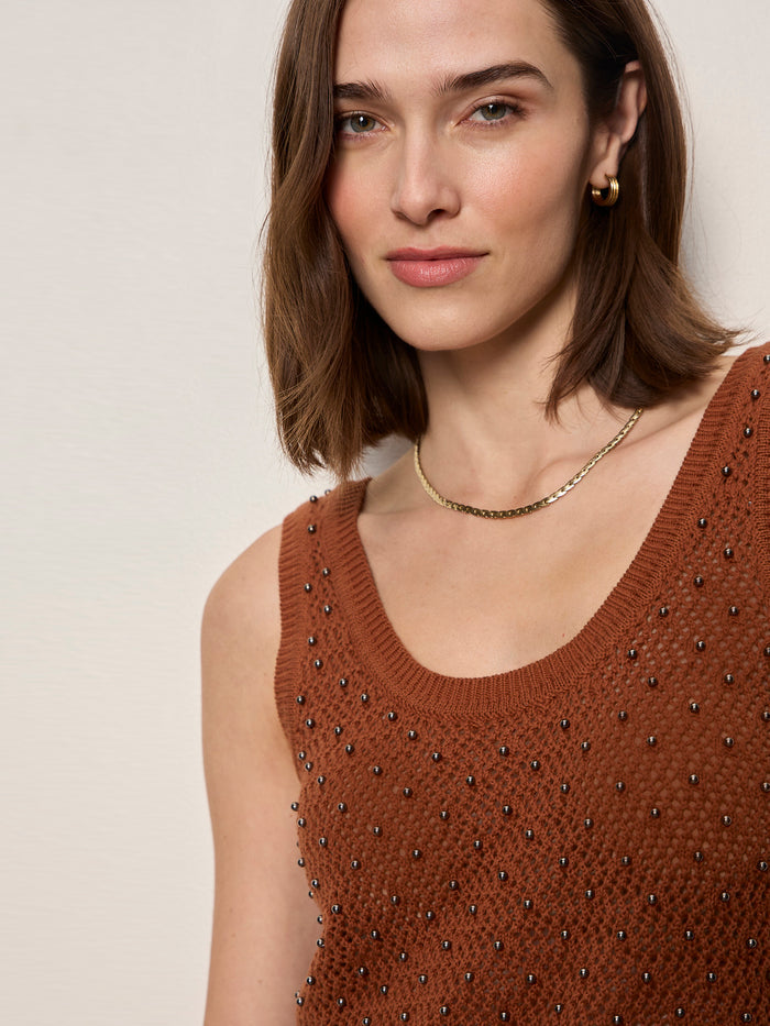 A woman with shoulder-length brown hair wears the Sanctuary Clothing dune studded tank in ancient clay, accessorized with a gold chain necklace and hoop earrings, standing against a light background and smiling at the camera.