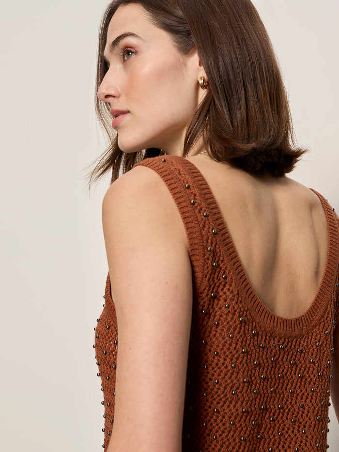 A woman with straight brown hair models the Sanctuary Clothing dune studded tank in ancient clay, a sleeveless, rust-colored knit top with bead details and a deep scoop back. She stands slightly turned away against a plain backdrop.