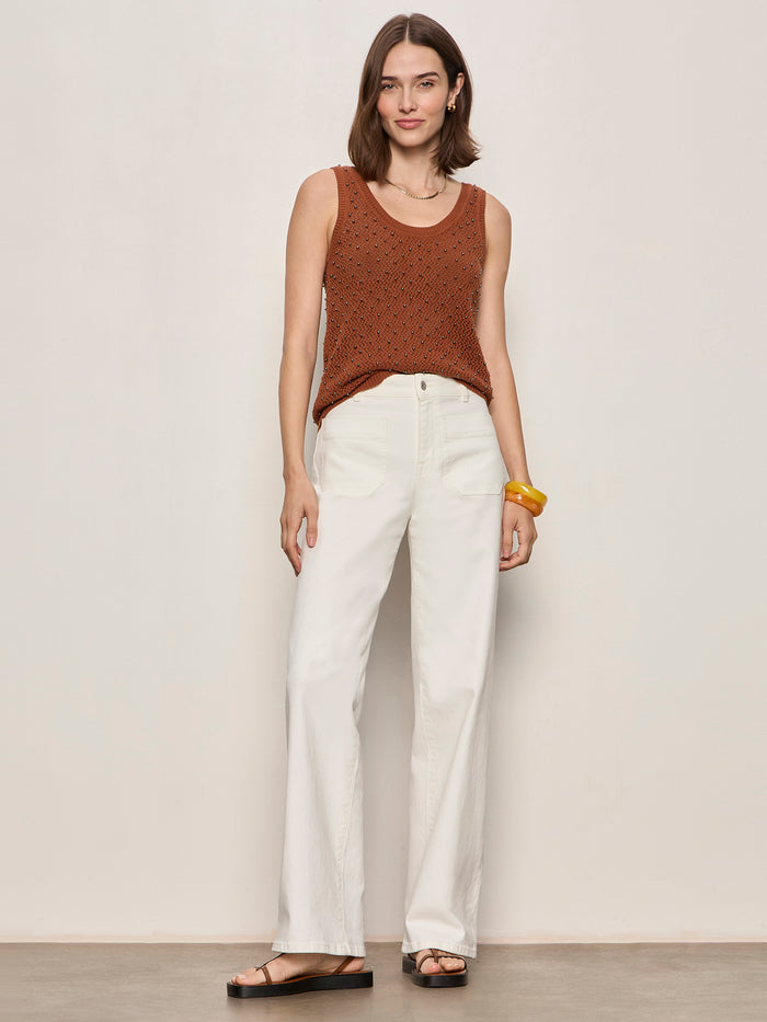 A woman with shoulder-length brown hair and a soft smile wears the Sanctuary Clothing dune studded tank in ancient clay, paired with white high-waisted wide-leg pants, black sandals, and a chunky yellow bracelet against a plain wall.