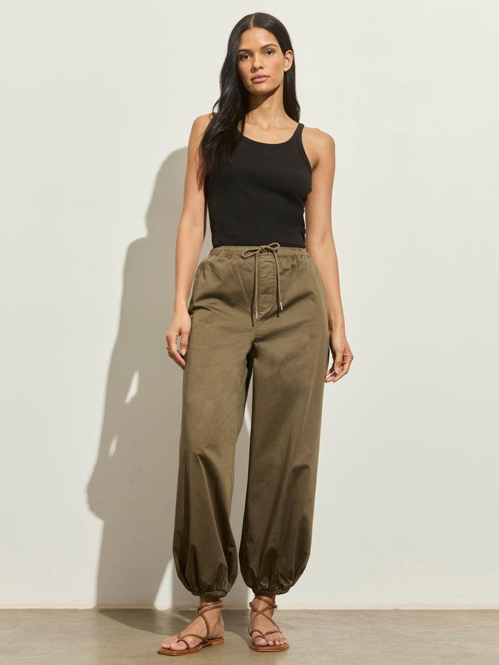 A woman with long dark hair and a relaxed expression stands against a light wall, wearing Sanctuary Clothing's float on balloon pant in timber, paired with a black sleeveless tank top and strappy flat sandals.