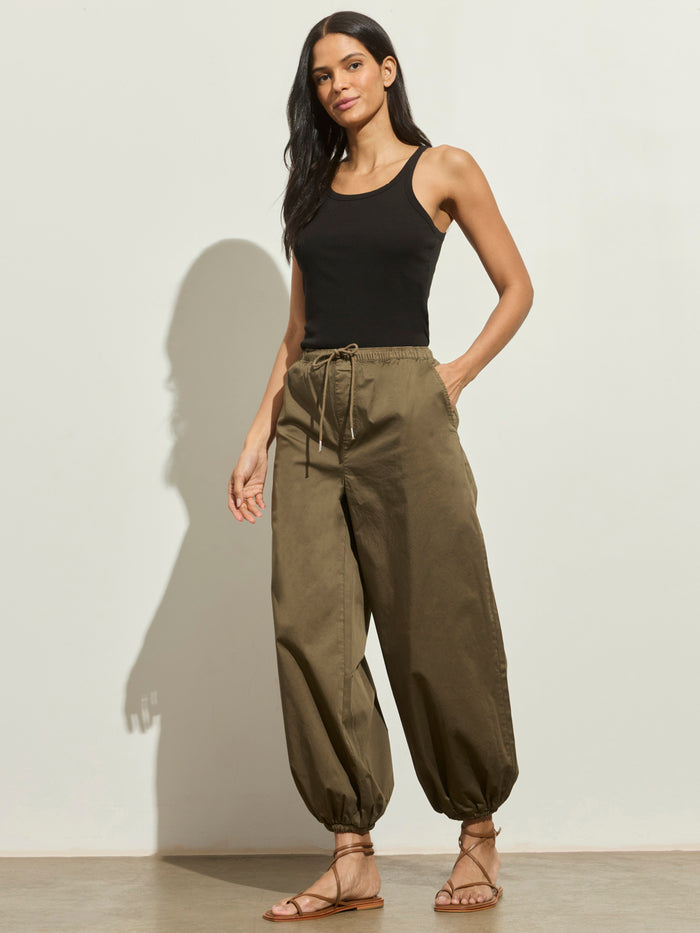 A woman with long dark hair smiles against a light background, wearing Sanctuary Clothing's "float on balloon pant timber"—olive green drawstring pants with elastic cuffs—paired with a black tank top and brown sandals, one hand in her pocket.