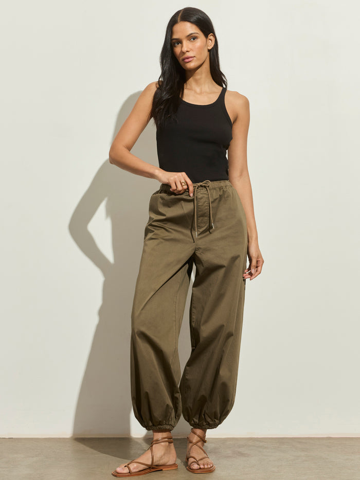 A woman with long dark hair and a relaxed pose stands against a light wall wearing Sanctuary Clothing’s float on balloon pant in timber, paired with a black sleeveless tank top and brown strappy sandals.