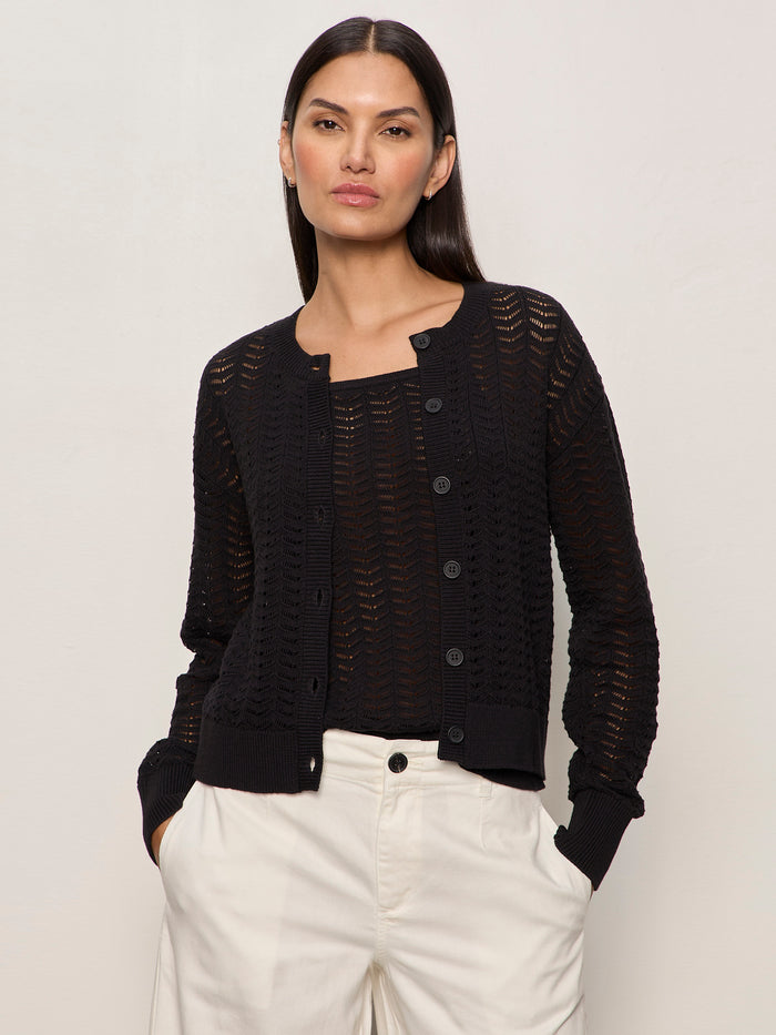 A woman with long dark hair models the Sanctuary Clothing "for keeps pointelle cardigan" in black over a matching top and light high-waisted trousers, standing with hands in pockets against a plain background.