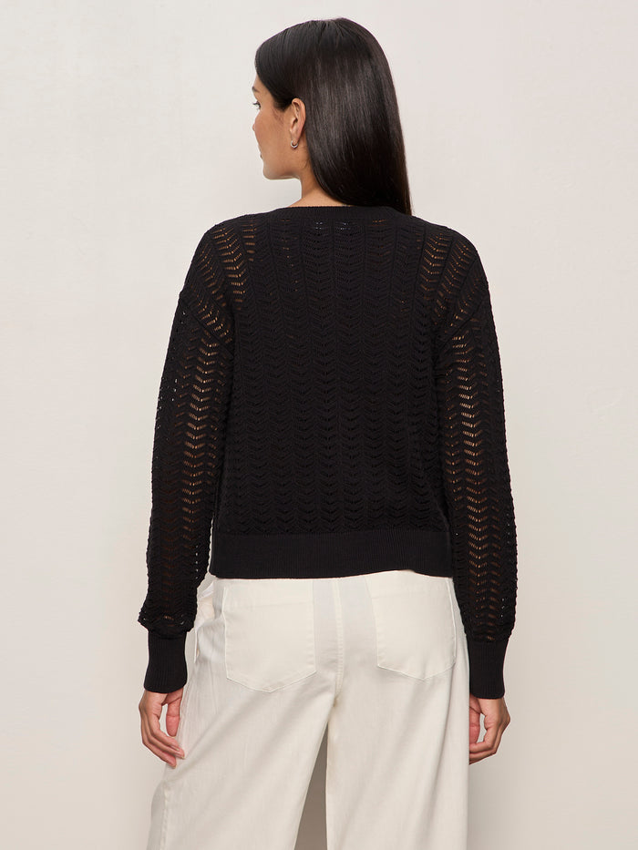 A woman with long dark hair, seen from behind, wears the Sanctuary Clothing "for keeps pointelle cardigan" in black with white pants, standing against a plain light-colored wall.