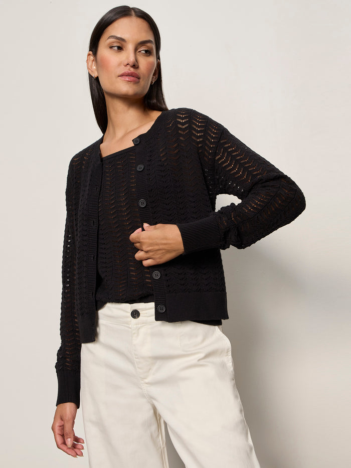 A woman wears the Sanctuary Clothing for keeps pointelle cardigan in black over a black top and white pants, standing against a plain light background and looking to the side with one hand on her waist.