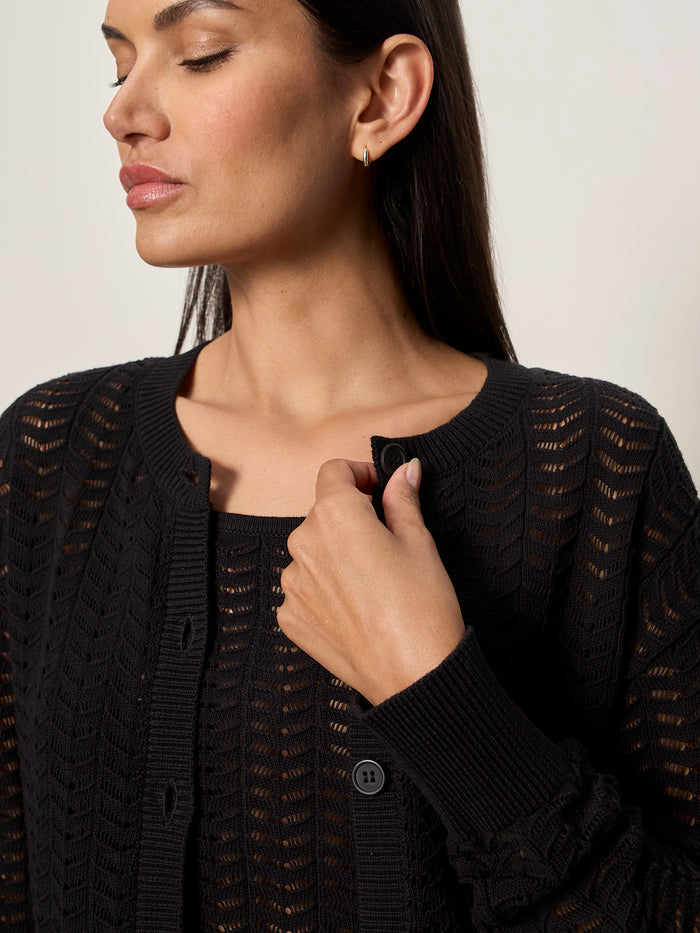 A woman with long dark hair wears the black "for keeps pointelle cardigan" by Sanctuary Clothing over a matching top. She faces left, gently holding the neckline, her expression calm and neutral.