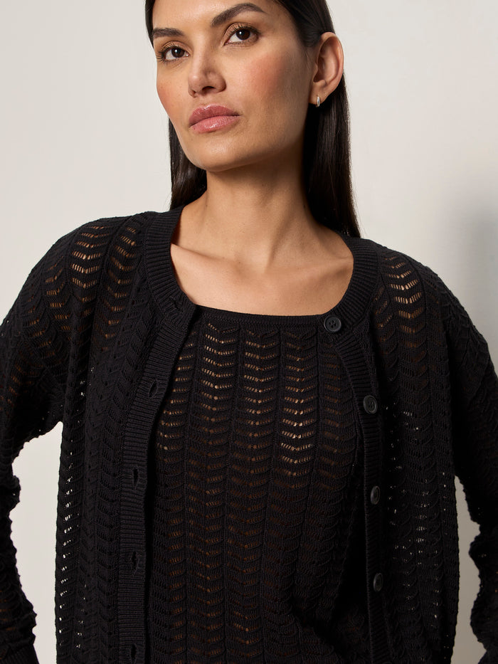 A woman with long dark hair wears the Sanctuary Clothing for keeps pointelle cardigan in black over a matching top, standing against a plain light background and looking slightly to the side.