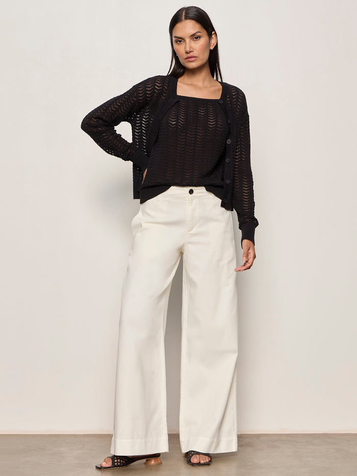 A woman poses with one hand on her hip, wearing the Sanctuary Clothing "for keeps pointelle cardigan" in black over a black top, white wide-leg pants, and black sandals, standing against a plain light wall with a neutral expression.