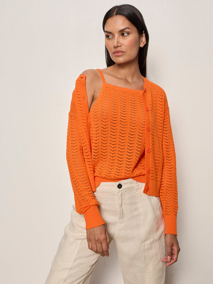 A woman wears the Sanctuary Clothing "for keeps pointelle cardigan" in hibiscus orange with high-waisted beige pants, posing against a plain light-colored wall.
