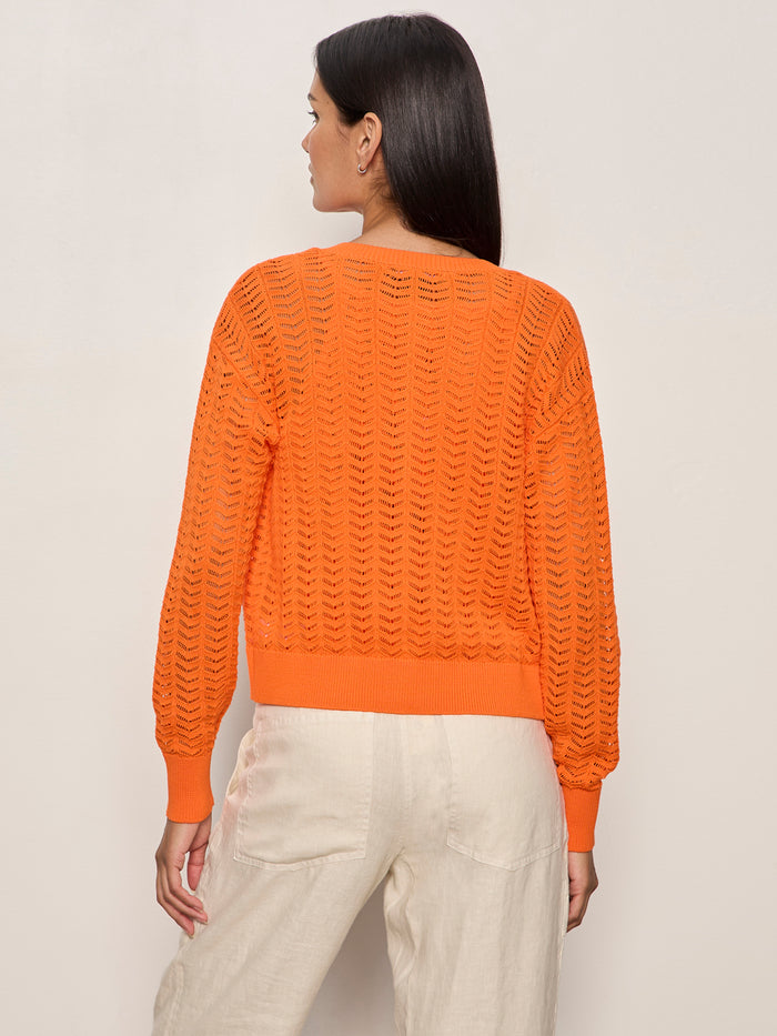 A woman faces away from the camera, wearing the Sanctuary Clothing "for keeps" pointelle cardigan in hibiscus orange with light beige pants, set against a plain light background.