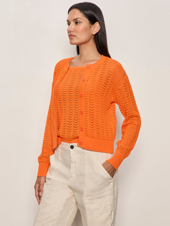 A woman with long dark hair stands against a light background wearing the Sanctuary Clothing "For Keeps" pointelle cardigan in hibiscus orange and light beige pants, her left hand tucked into her pocket and a neutral expression on her face.