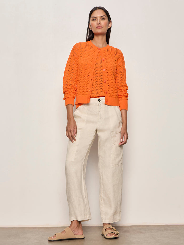 A woman with straight dark hair wears the Sanctuary Clothing for keeps pointelle cardigan in hibiscus orange, paired with off-white pants and beige slide sandals, standing against a plain light wall with minimal jewelry and a neutral expression.