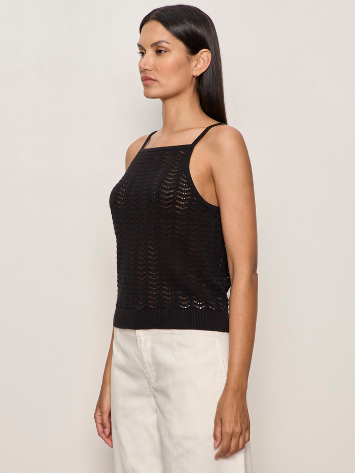 A woman stands in profile against a light background, wearing the Sanctuary Clothing "for keeps pointelle tank" in black with thin straps and off-white trousers. Her straight dark hair is worn down, and she has a neutral expression.