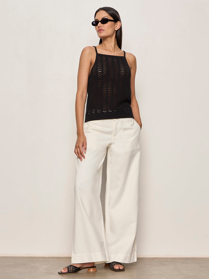 A woman stands against a light wall wearing the Sanctuary Clothing for keeps pointelle tank in black, black sunglasses, wide-leg white pants, and black sandals. Her hands are in her pockets as she looks to the side.