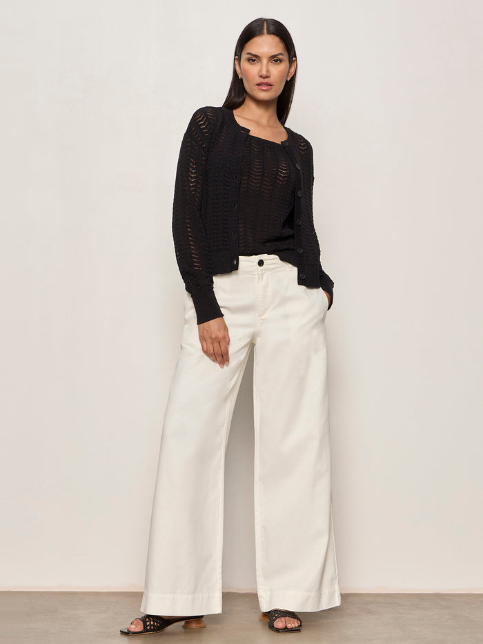 A woman with straight dark hair stands against a plain wall wearing the Sanctuary Clothing for keeps pointelle tank in black, wide-leg white pants, and black sandals. She poses relaxed with one hand in her pocket and a neutral expression.
