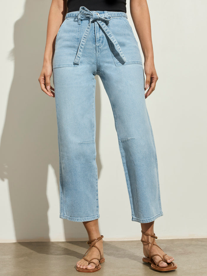 A person stands against a light wall, wearing Sanctuary Clothing’s foundry jean sailor blue with a tie belt. The photo is cropped to show only the lower torso and legs, paired with a black top and tan strappy sandals.