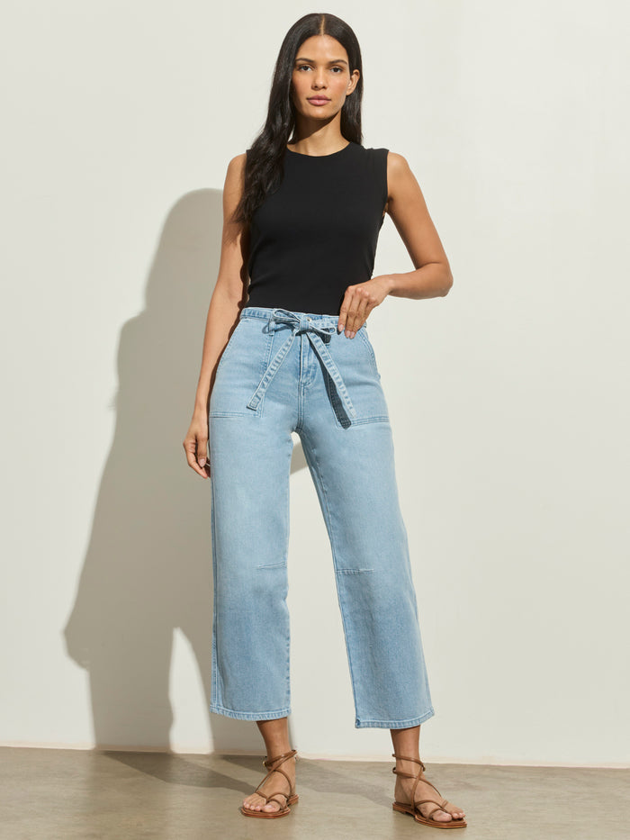 A woman with long dark hair stands confidently by a light wall, wearing Sanctuary Clothing's foundry jean sailor blue—high-waisted, wide-leg jeans with a belt—paired with a sleeveless black top and brown strappy sandals.