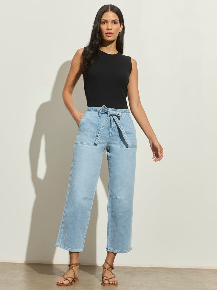 A woman with long dark hair stands against a plain wall, wearing the Sanctuary Clothing foundry jean sailor blue—light blue high-waisted jeans with a tied belt—paired with a sleeveless black top and tan strappy sandals, hands in her pockets.