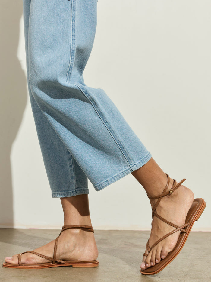 A person models the Sanctuary Clothing foundry jean in sailor blue, paired with brown strappy sandals, standing on a neutral floor with one foot slightly raised against a plain light background.